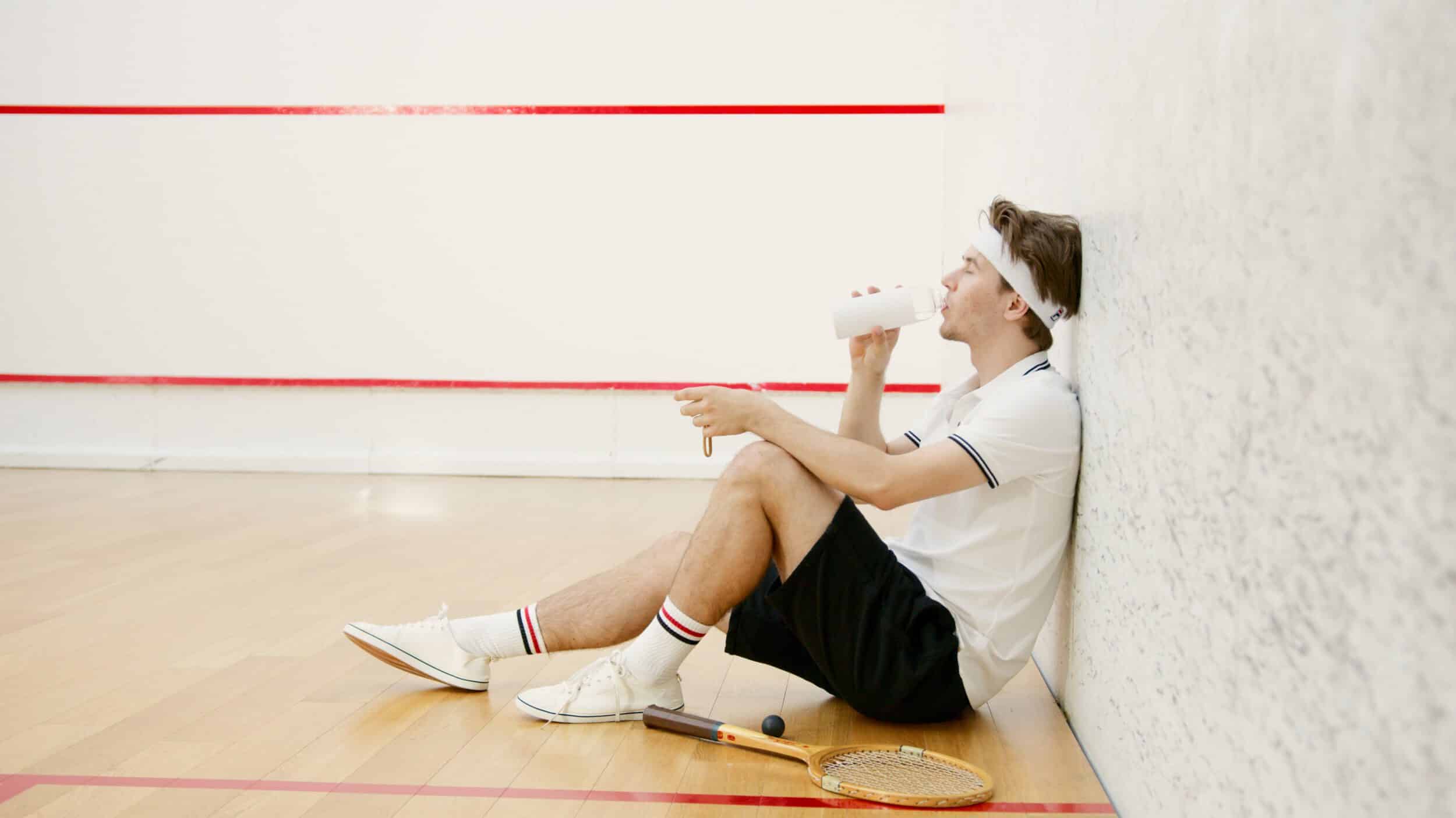 Tennisspieler, der auf dem Boden sitzt und Wasser aus einer Trinkflasche trinkt.